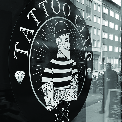 En svartvit fönsterdekla till en tatueringsstudio. Den är rund med en sjömans överkropp i mitten. Ovanför står det Tattoo club i en båge som följer formen på dekalen.